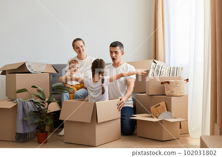 Horizontal shot of happy delighted family unpacking moving box at their new home and playing with cardboard boxes, couple with their kids having fun while moving in new flat. Horizontal shot of happy delighted family unpacking moving box at their new home and playing with cardboard boxes, couple with their kids having fun while moving in new flat. 102031802