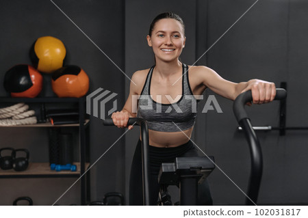 Portrait of smiling fitness woman using air bike for cardio workout and exercising her legs at gym 102031817