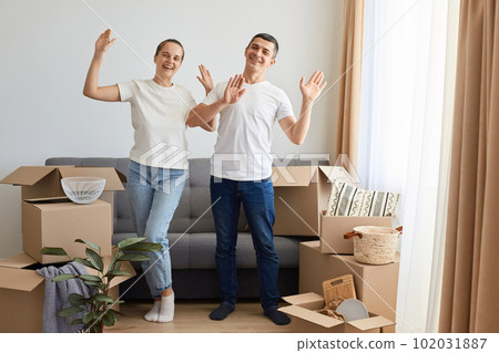 Portrait of extremely happy woman and man wearing jeans and white t shirts posing among cardboard boxes and dancing, celebrating buying new their own apartment. 102031887