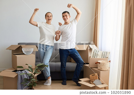 Full length portrait of satisfied woman and man wearing jeans and white t shirts posing among their belongings in boxes, standing with raised arms, wife and husband celebrating relocation, dancing. Full length portrait of satisfied woman and man wearing jeans and white t shirts posing among their belongings in boxes, standing with raised arms, wife and husband celebrating relocation, dancing. 102031890