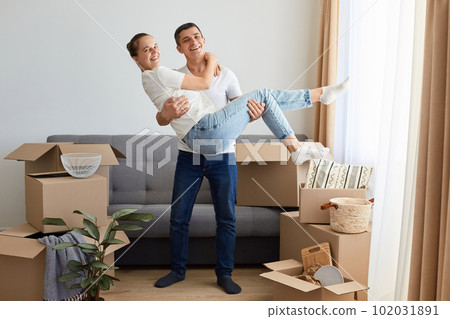 Full length portrait of loving couple in a new house, woman and man wearing jeans and white t shirts posing among their personal piles in carton parcels, husband holding wife in hands. 102031891