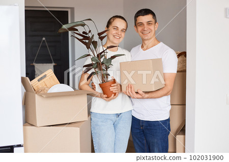 Indoor shot of happy couple wearing casual style clothing standing with personal pile and flower, looking at camera with charming smiles, relocating, moving. Indoor shot of happy couple wearing casual style clothing standing with personal pile and flower, looking at camera with charming smiles, relocating, moving. 102031900