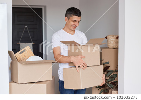 Portrait of optimistic handsome man wearing white T-shirt standing with cardboard boxes with personal stuff, looking inside with happy expression, relocation, real estate. Portrait of optimistic handsome man wearing white T-shirt standing with cardboard boxes with personal stuff, looking inside with happy expression, relocation, real estate. 102032273