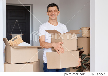 Horizontal shot of smiling man wearing white t shirt standing with cardboard box in hands, looking at camera with smile, being happy to move into a new house. Horizontal shot of smiling man wearing white t shirt standing with cardboard box in hands, looking at camera with smile, being happy to move into a new house. 102032274