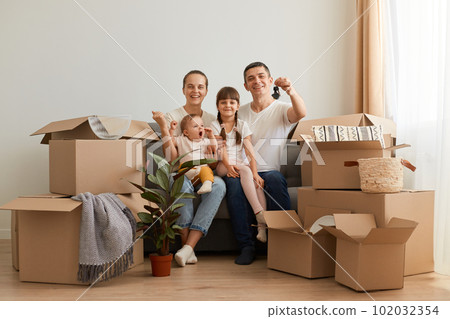 Portrait of extremely happy couple sitting on sofa with children after moving, buying new apartment, showing keys of house they are relocating, surrounded with cardboard boxes. 102032354