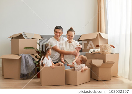 Indoor shot of happy lovinng couple wearing casual style white t shirts posing with carton boxes during moving to a new flat, hugging each other and looking at their daughters in boxes with smile. Indoor shot of happy lovinng couple wearing casual style white t shirts posing with carton boxes during moving to a new flat, hugging each other and looking at their daughters in boxes with smile. 102032397