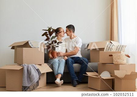 Horizontal shot of lovely romantic young family buying new flat and posing surrounded with boxes with personal pile, sitting on sofa and kissing, celebrating relocation into a new flat. Horizontal shot of lovely romantic young family buying new flat and posing surrounded with boxes with personal pile, sitting on sofa and kissing, celebrating relocation into a new flat. 102032511