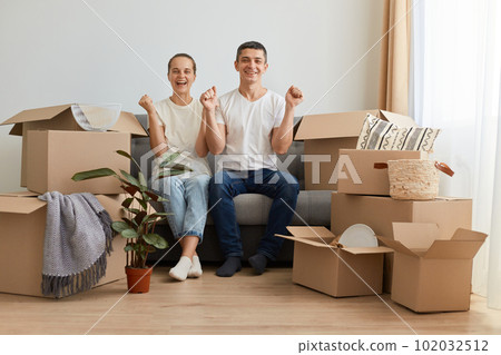 Indoor shot of happy optimistic delighted young couple sitting on sofa surrounded with carton boxes with personal belongings, clenched fists, celebrating buying new house. Indoor shot of happy optimistic delighted young couple sitting on sofa surrounded with carton boxes with personal belongings, clenched fists, celebrating buying new house. 102032512