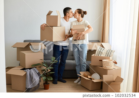 Romantic young couple posing near sofa surrounded with carton boxes, family during relocation to their own house, standing with parcels and kissing each other. 102032534