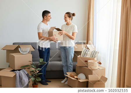 Indoor shot of positive pleasant young couple standing near sofa surrounded with carton boxes, family during relocation, expressing positive emotions and happiness. 102032535