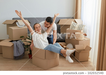 Happy couple having fun laughing moving into new home, young excited woman riding sitting in cardboard box while man pushing it, cheerful family playing while packing and unpacking belongings together Happy couple having fun laughing moving into new home, young excited woman riding sitting in cardboard box while man pushing it, cheerful family playing while packing and unpacking belongings together 102032540