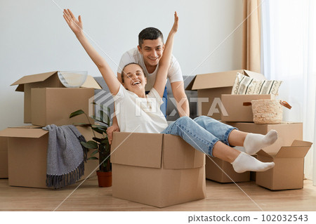Happy couple having fun while moving into new house, excited woman with raised arms riding sitting in cardboard box while man pushing it, cheerful family playing while unpacking belongings together. Happy couple having fun while moving into new house, excited woman with raised arms riding sitting in cardboard box while man pushing it, cheerful family playing while unpacking belongings together. 102032543
