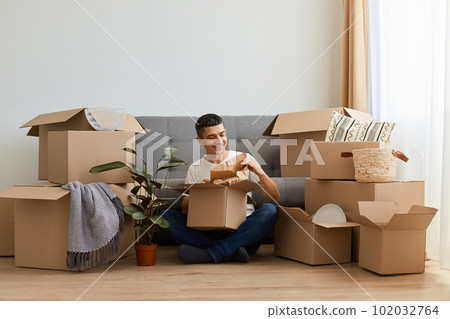 Horizontal shot of brunette positive man wearing white t shirt sitting on floor near sofa surrounded with cardboard boxes, upacking his personal piles after moving in. Horizontal shot of brunette positive man wearing white t shirt sitting on floor near sofa surrounded with cardboard boxes, upacking his personal piles after moving in. 102032764