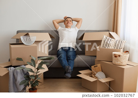 Happy satisfied relaxed man wearing white T-shirt and jeans sitting on sofa surrounded with pile of packages, looking at camera with smile, keeping hands behind head, being glad to move to a new flat. 102032767