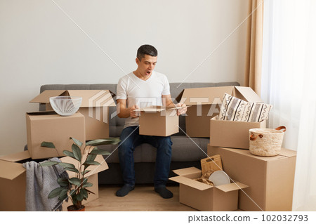 Indoor shot of brunette man wearing white casual T-shirt and jeans, sitting on sofa, unpacking boxes after relocating, looking inside box with shocked facial expression. Indoor shot of brunette man wearing white casual T-shirt and jeans, sitting on sofa, unpacking boxes after relocating, looking inside box with shocked facial expression. 102032793