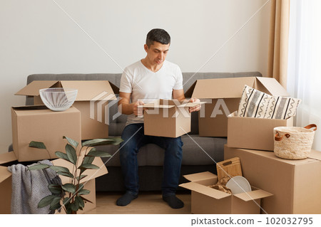 Sad man wearing white T-shirt and jeans sitting on sofa surrounded with cardboard boxes, posing with pout lips and sorrow expression, looking inside package, upset of broken things after moving. Sad man wearing white T-shirt and jeans sitting on sofa surrounded with cardboard boxes, posing with pout lips and sorrow expression, looking inside package, upset of broken things after moving. 102032795