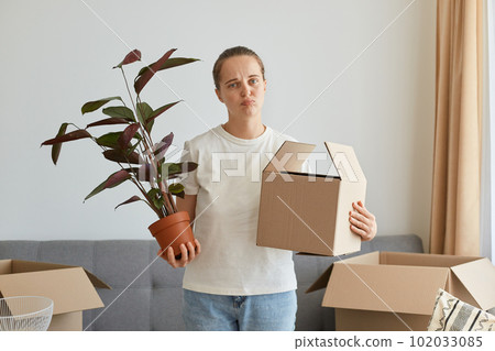 Portrait of sad upset woman wearing white t shirt standing with cardboard box, relocating to a new house, packing personal pile, feeling sorrow to left her old house and moving to another flat. Portrait of sad upset woman wearing white t shirt standing with cardboard box, relocating to a new house, packing personal pile, feeling sorrow to left her old house and moving to another flat. 102033085