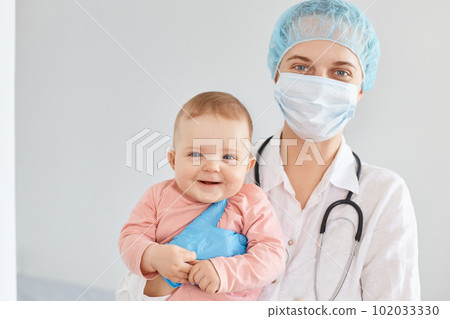 Indoor shot of beautiful woman family doctor wearing medical uniform and surgical mask holding infant baby in hands, children health care, visit to the pediatrician. 102033330