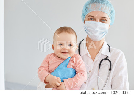 Winsome female doctor pediatrician wearing medical cap, gloves and protective mask, standing with toddler baby girl in hands, little child at the clinic or hospital. 102033331