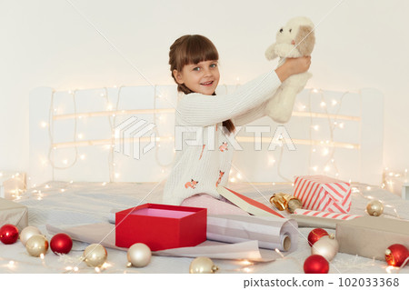Side view portrait of little girl wearing white sweater holding soft toy dog, sitting on bed with Christmas decoration and garland, raised arms and showing her New year present. 102033368