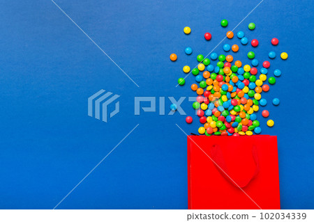 shopping paper gift bag in corner full of assorted traditional candies falling out on colored background with copy space. Happy Holidays sale concept shopping paper gift bag in corner full of assorted traditional candies falling out on colored background with copy space. Happy Holidays sale concept 102034339