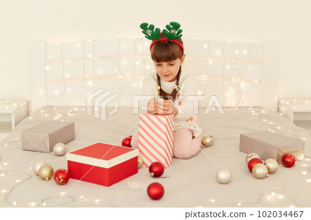 Indoor shot of pretty little girl wearing white sweater and deer horns carnival hoop, sitting on bed with Christmas presents, holding baubles for x mas tree decoration in hands. Indoor shot of pretty little girl wearing white sweater and deer horns carnival hoop, sitting on bed with Christmas presents, holding baubles for x mas tree decoration in hands. 102034467