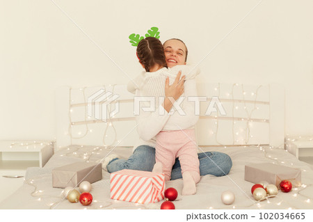 Indoor shot of happy mother and her daughter wearing white sweaters hugging each other while sitting on bed with Christmas decoration, happy new year. Indoor shot of happy mother and her daughter wearing white sweaters hugging each other while sitting on bed with Christmas decoration, happy new year. 102034665