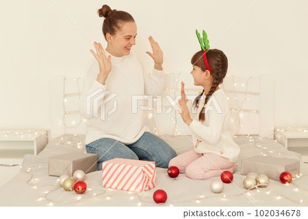 Indoor shot of optimistic mother and daughter wearing casual style white sweaters sitting on bed, giving five to each other, celebrating Christmas, happy new year. 102034678