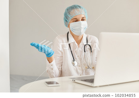 Confused female doctor wearing medical cap, surgical mask, rubber gloves and gown, using notebook for working, looking at screen, having video call, spreading hands aside, showing helpless gesture. Confused female doctor wearing medical cap, surgical mask, rubber gloves and gown, using notebook for working, looking at screen, having video call, spreading hands aside, showing helpless gesture. 102034855