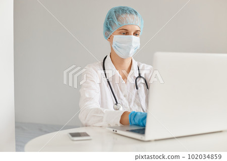 Beautiful female doctor wearing medical cap, surgical mask, rubber gloves and gown, using notebook for working, typing on keyboard, writing new medicine scientific article. Beautiful female doctor wearing medical cap, surgical mask, rubber gloves and gown, using notebook for working, typing on keyboard, writing new medicine scientific article. 102034859