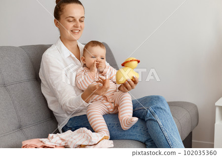 Smiling happy caucasian woman wearing white shirt and jeans, sitting on sofa with her toddler daughter, mommy playing with kid, showing yellow toy for her, expressing happiness. 102035360