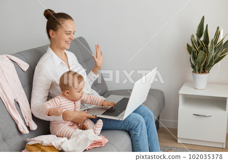 Side view profile of smiling happy woman wearing white shirt and jeans sitting on sofa with baby daughter and holding laptop, waving hand to display, saying hello or bye in video call. 102035378