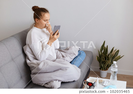 Profile portrait of sick female wearing white sweater sitting on sofa wrapped in blanket, holding phone in hands, checking social networks while staying home during virus illness. Profile portrait of sick female wearing white sweater sitting on sofa wrapped in blanket, holding phone in hands, checking social networks while staying home during virus illness. 102035465