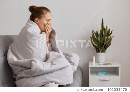 Sick young adult woman with hair bun sitting on sofa wrapped in blanket, having symptoms of pneumonia and bronchitis, coughing, suffering headache and high temperature. Sick young adult woman with hair bun sitting on sofa wrapped in blanket, having symptoms of pneumonia and bronchitis, coughing, suffering headache and high temperature. 102035476