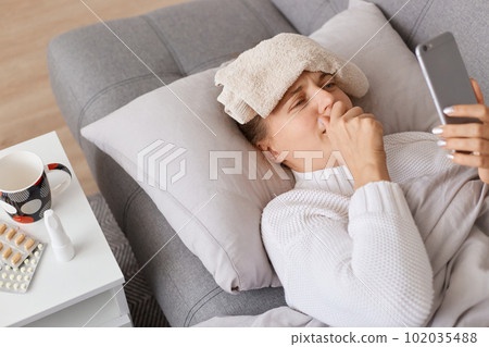 Portrait of sick unhealthy girl staying at home on sofa and uses smartphone to talk to her doctor via video conference, medical application, wearing white sweater posing with wet towel on forehead. Portrait of sick unhealthy girl staying at home on sofa and uses smartphone to talk to her doctor via video conference, medical application, wearing white sweater posing with wet towel on forehead. 102035488
