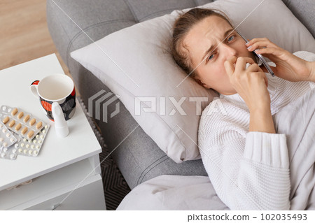 Portrait of sad sick woman lying on sofa on pillow, looking at camera with upset expression, using napkin for runny nose, talking with doctor asking about treatment. Portrait of sad sick woman lying on sofa on pillow, looking at camera with upset expression, using napkin for runny nose, talking with doctor asking about treatment. 102035493