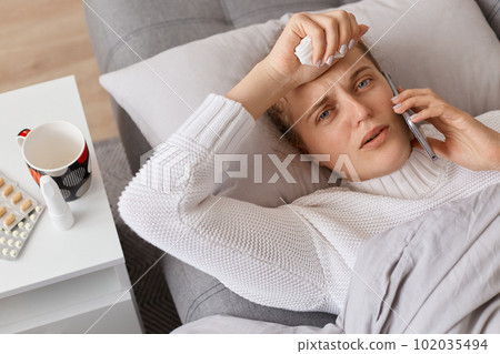 Indoor shot of sick girl lying in bed and uses smartphone to talk to her doctor, suffering headache and high temperature, looking at camera, expressing sorrow. 102035494