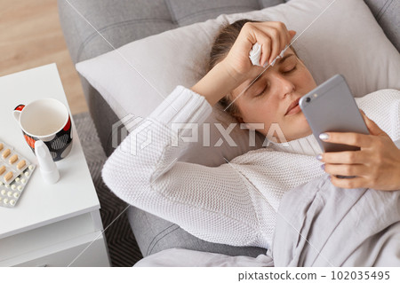 Indoor shot of sick sad female wearing white sweater lying on sofa and using smart phone, dialing doctor number or searching information about grippe treatment. Indoor shot of sick sad female wearing white sweater lying on sofa and using smart phone, dialing doctor number or searching information about grippe treatment. 102035495