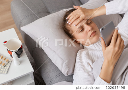 Portrait of unhealthy with lying on sofa and touching her forehead, having terrible headache, calling doctor, reading pills instruction online, searching information about her sickness in Internet. Portrait of unhealthy with lying on sofa and touching her forehead, having terrible headache, calling doctor, reading pills instruction online, searching information about her sickness in Internet. 102035496