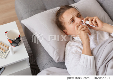 Portrait of sick woman lying on sofa with high fever, catching cold and flu, using nasal spray for runny nose, expressing upset emotions, suffering grippe or influenza. Portrait of sick woman lying on sofa with high fever, catching cold and flu, using nasal spray for runny nose, expressing upset emotions, suffering grippe or influenza. 102035497