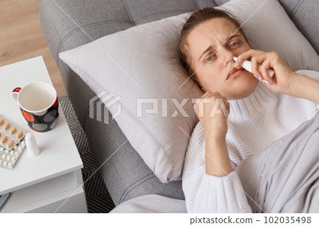 Unhealthy ill woman lying on sofa and using nasal spray, looking at camera, treating grippe, staying at home, having influenza symptoms, need treatment. Unhealthy ill woman lying on sofa and using nasal spray, looking at camera, treating grippe, staying at home, having influenza symptoms, need treatment. 102035498