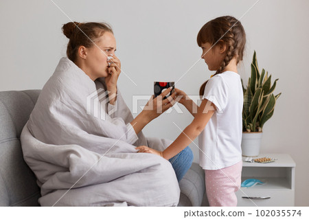 Side view portrait of caring child bringing hot tea for her sick mother suffering influenza or pneumonia, posing with napkin, having runny nose and high temperature. Side view portrait of caring child bringing hot tea for her sick mother suffering influenza or pneumonia, posing with napkin, having runny nose and high temperature. 102035574