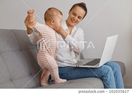 Delighted smiling dark haired woman spending time with toddler daughter and working on notebook, sitting on sofa in living room, expressing positive emotions. 102035627
