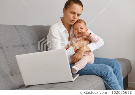 Portrait of unhappy sad woman with hair bun wearing white shirt and jeans sitting on cough with her crying toddler daughter, trying to calm down infant baby, hugging child and hush. 102035638