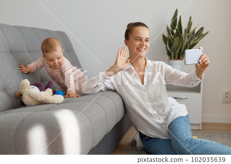 Indoor shot of friendly woman wearing white shirt and jeans sitting on floor and having video call with somebody while her infant daughter playing on sofa. 102035639