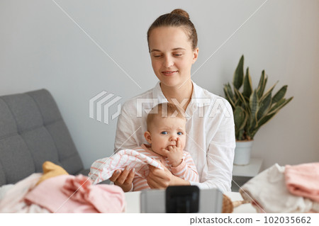 Beautiful young adult woman blogger sitting in front of phone on tripod and recording video together with her toddler child, posing at table and being surrounded with baby clothing after online Beautiful young adult woman blogger sitting in front of phone on tripod and recording video together with her toddler child, posing at table and being surrounded with baby clothing after online 102035662
