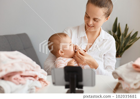 Indoor shot of optimistic Caucasian mother sitting at table with toddler baby, giving nipple to her little kid, using mobile phone on tripod for broadcasting livestream. Indoor shot of optimistic Caucasian mother sitting at table with toddler baby, giving nipple to her little kid, using mobile phone on tripod for broadcasting livestream. 102035681
