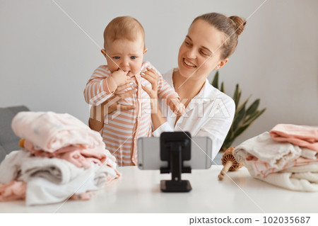 Attractive smiling young female with hair bun broadcasting livestream at home together with her infant daughter, talking with subscribers, filming content for her blog. Attractive smiling young female with hair bun broadcasting livestream at home together with her infant daughter, talking with subscribers, filming content for her blog. 102035687