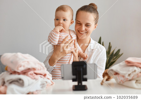 Horizontal shot of satisfied woman blogger sitting at table with toddler kid and recording video for her vlog, making content with her daughter, having pleasant conversation with subscribers. Horizontal shot of satisfied woman blogger sitting at table with toddler kid and recording video for her vlog, making content with her daughter, having pleasant conversation with subscribers. 102035689
