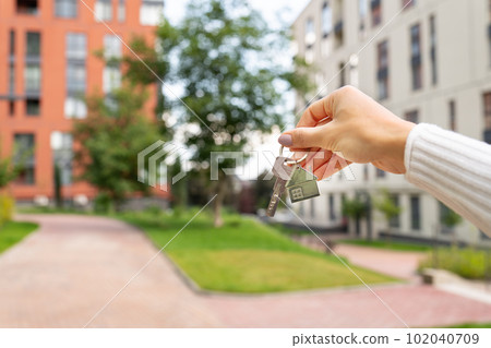 Hand holding keys on the background of a new building, construction concept. Buying real estate, housing, apartments. Private yard, park, kindergarten, playgrounds. 102040709